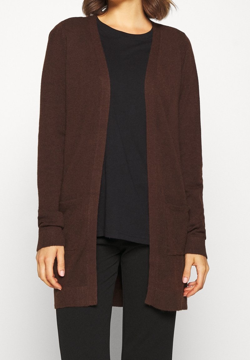 Femme portant un cardigan marron foncé ouvert sur le devant avec des manches longues et des poches avant, par-dessus une chemise noire et un pantalon noir.