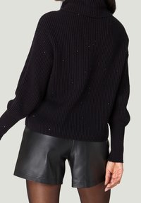 Mujer vestida con un suéter negro de canalé con pequeñas lentejuelas y pantalones cortos de cuero negro, mostrada desde atrás contra un fondo neutro.