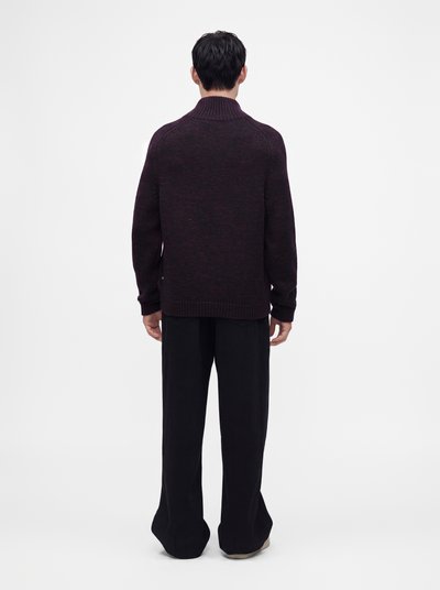 Homme aux cheveux foncés portant un pull en maille violet foncé et un pantalon noir ample, debout de dos sur un fond blanc.