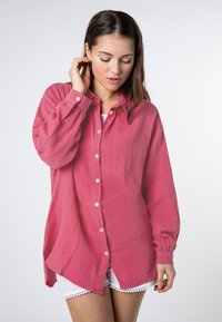 Roze overhemd met knoopsluiting van textuurstof, met lange mouwen, een gebogen zoom en witte knopen. Draagt over witte shorts met kantafwerking.