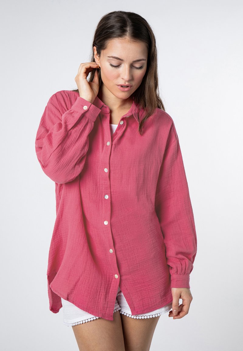 Rosa Hemdbluse aus strukturiertem Stoff mit langen Ärmeln, leicht abfallendem Saum und weißen Knöpfen, kombiniert mit weißen Shorts.