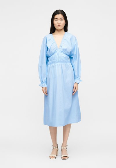 Γυναίκα με ανοιχτού μπλε midi φόρεμα με μακριά μανίκια, βαθύ V-λαιμόκοψη και ζώνη στη μέση, συνδυασμένο με μπεζ πέδιλα με λουράκια, στέκεται μπροστά σε λευκό φόντο.