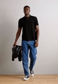 Schwarzes Poloshirt, blaue Jeans und schwarze Sneakers. Hält eine schwarze Lederjacke. Steht auf einem Holzboden vor einem schlichten Hintergrund.