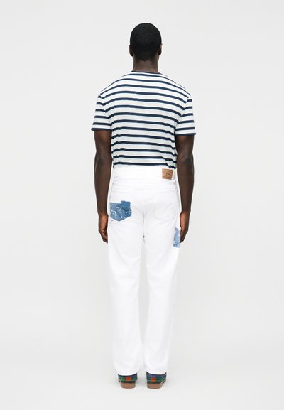 Άνδρας που φοράει λευκά τζιν με μπλε μπαλώματα στις πίσω τσέπες και μπλε και λευκή ριγέ κοντομάνικη μπλούζα, όρθιος με την πλάτη στραμμένη.