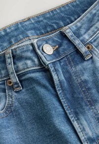 Mėlynos džinsinės kelnės su klasikiniu penkių kišenių dizainu, metaliniu mygtuko užsegimu ir matomomis siūlėmis. Audinys šiek tiek išblukęs.