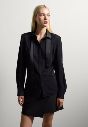 Camicia nera a maniche lunghe con dettaglio in cravatta di satin, con bottoni sul davanti e polsini. Abbinata a una gonna nera elegante, entrambe in tessuto liscio.