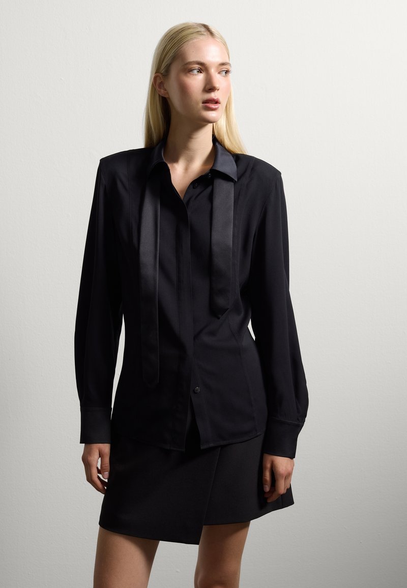 Black long-sleeve shirt with a satin tie detail, button front, and cuffs. Paired with a sleek black skirt, both in smooth fabric.