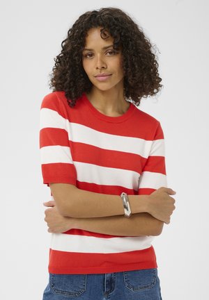 Femme aux cheveux bouclés portant un pull à manches courtes rouge et blanc rayé, un jean bleu et un bracelet argenté, debout les bras croisés.