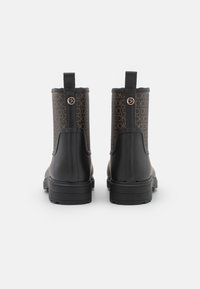 Une paire de bottes noires à la cheville avec une tige marron à motifs comportant le logo "CK" répété, vue de l'arrière sur un fond blanc.
