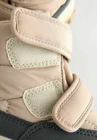 Beige og grå fodtøj med et tekstureret stofoverdel og velcrolukninger. Bemærkelsesværdige syninger og en polstret krave for komfort.