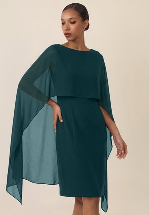 Mujer con vestido verde oscuro hasta la rodilla y mangas de capa translúcidas, de pie con una mano en la cadera contra un fondo neutro.