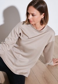 Maglietta a maniche lunghe color beige chiaro realizzata in tessuto morbido, con scollo rotondo e vestibilità comoda, arricchita da dettagli di cuciture sottili.