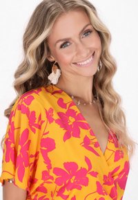 Helles gelbes Kleid mit großem pinkem Blumenmuster und kurzen, lockeren Ärmeln; accessorisiert mit einer Perlenkette und Federohrringen.
