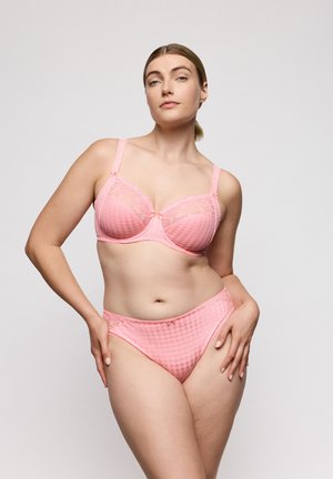 Vrouw draagt bijpassende roze kanten beha en slipje, staat met één hand op de heup tegen een effen lichte achtergrond.