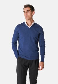 Maglione a V in tessuto a maglia blu scuro uniforme, con maniche lunghe e polsini a coste. Indossato sopra una camicia bianca con colletto, abbinato a pantaloni neri.