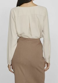 Donna che indossa una blusa lunga beige con maniche lunghe, infilata in una gonna marrone a vita alta, vista di spalle su uno sfondo neutro.