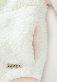 Giacca in pile bianca con una superficie morbida e strutturata, con un polsino beige e un'etichetta metallica con la scritta "BAKER" attaccata alla manica.
