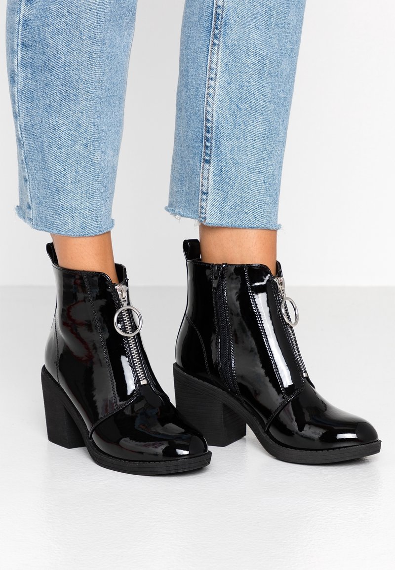 Jambes portant un jean bleu clair raccourci et des bottines noires brillantes à talons épais avec une fermeture éclair à l'avant munie d'une tirette circulaire.