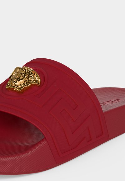 Sandale slide rouge avec motif géométrique embossé et emblème tête de Méduse doré sur la bride.
