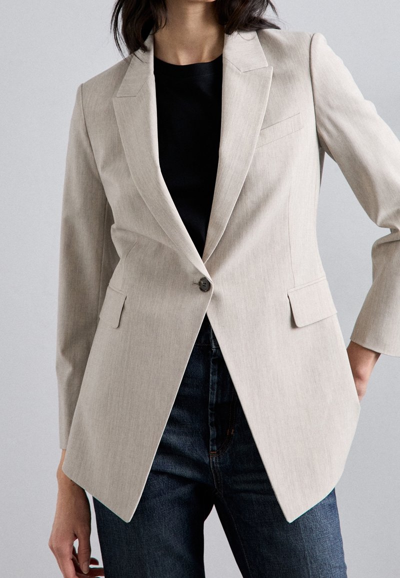 Kobieta ubrana w beżową marynarkę z jednym guzikiem, czarny top i ciemnoniebieskie dżinsy, pozująca z jedną ręką na biodrze na jednolitym tle.