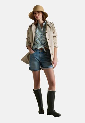 Sieviete, kas valkā bēša krāsas tranšeju mēteli, ziedu raksta šalli, džinsa šortus, melnus gumijas zābakus un plaša malu salmu cepuri, stāvot ar vienu roku kabatā.