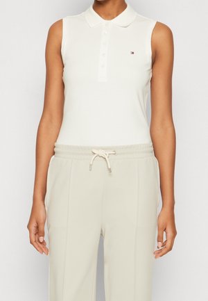 Vrouw die een mouwloze witte poloshirt met knopen draagt en beige broek met een trekkoord in de taille, staand tegen een effen achtergrond.