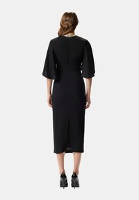 Vestito midi nero con maniche a campana, cerniera posteriore e spacco. Presenta una scollatura tonda decorata con sottili ornamenti. Indossato con tacchi neri.