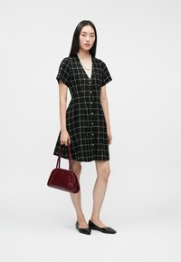 Čierne kárované šaty s krátkymi rukávmi, výstrihom do V, zlatými gombíkmi, v kombinácii s červenou kabelkou a čiernymi špicatými balerínkami. Hladký materiál.