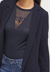Veste de blazer marine portée sur un haut côtelé marine avec un motif en dentelle au niveau de l'encolure. L'ensemble est associé à un jean denim bleu clair.