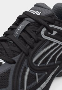 Κοντινή λήψη μαύρου παπουτσιού τρεξίματος Puma Skyet με μαύρα κορδόνια, υφή από δίχτυ και ανακλαστικά ασημένια στοιχεία στις πλαϊνές επιφάνειες.