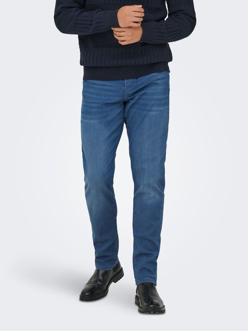 Uomo con jeans slim fit blu, maglione nero e stivali di pelle neri, in piedi davanti a uno sfondo semplice.