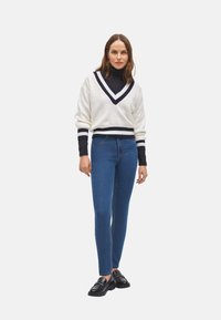 Suéter blanco de punto con cuello en V azul marino y rayas; combinado con jeans ajustados azules y mocasines negros. Textura suave, corte relajado.