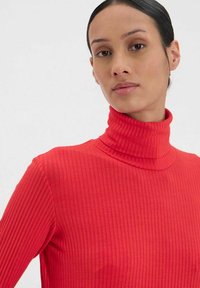 Pull en maille côtelée rouge avec un design ajusté. Il présente un col montant et un tissu texturé. La vue en gros plan met en avant les coutures et l'ajustement.