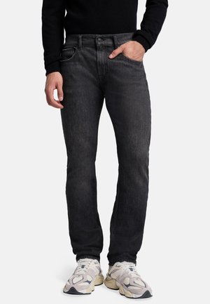 Jeans in denim neri con taglio dritto, leggera scoloritura, hardware minimo e cinque tasche, abbinati a scarpe da ginnastica di colore chiaro.