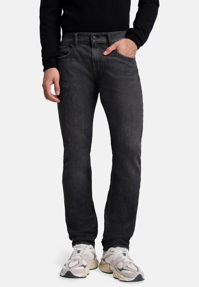 Schwarze Denim-Jeans mit geradem Schnitt, leichter Ausbleichung, minimalem Hardware-Einsatz und fünf Taschen, kombiniert mit hellen Sportschuhen.