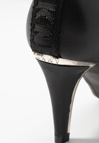 Svarta högklackade skor med en slät läderfinish, texturerad rem och metallisk detalj på hälen med ingraverad varumärkesinformation.