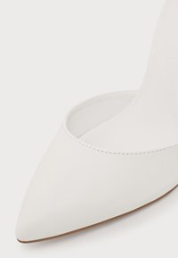 Gros plan sur une chaussure blanche à bout pointu avec une texture lisse et des coutures visibles le long du bord, montrant une partie de la semelle.