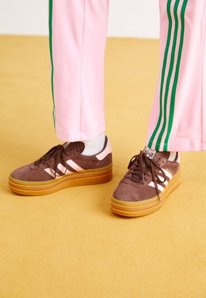 Osoba nosząca brązowe sneakersy Adidas Gazelle z gumowymi podeszwami, białe skarpetki i różowe spodnie z zielonymi paskami stoi na żółtej podłodze.