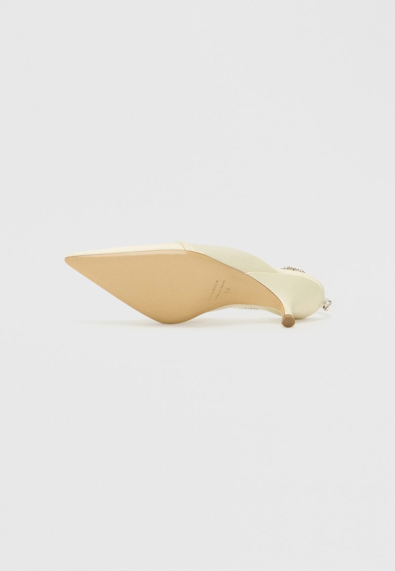 Zapato de tacón alto con punta afilada, hecho de material de color crema, con un tacón dorado claro y un acento decorativo en la parte trasera.