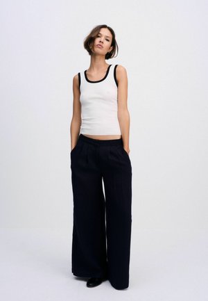 Donna che indossa un top bianco senza maniche con bordatura nera e pantaloni neri a gamba larga, in piedi con le mani nelle tasche su uno sfondo semplice.