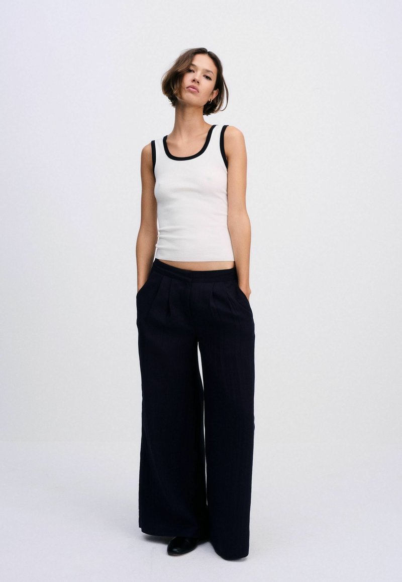 Femme portant un haut blanc sans manches avec bordure noire et un pantalon noir à jambes larges, debout les mains dans les poches devant un fond uni.