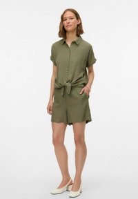 Romper corto olive green con maniche corte e bottoni, con un cintura annodata, caratterizzato da una vestibilità comoda e una texture liscia. Indossato con scarpe di colore chiaro.