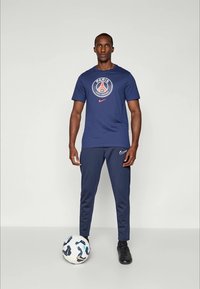 Nike sportsoutfit bestående af en marineblå t-shirt med et Paris-logo og matchende joggers. Sorte træningssko og en mønstret fodbold er inkluderet.