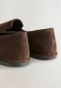 Chaussures slip-on en daim marron avec semelles noires, coutures visibles le long des bords, photographiées de dos sur une surface blanche.