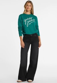 Tirkizna majica dugih rukava s bijelim tekstom "guess jeans", uparena s crnim širokim hlačama visokog struka. Ogrnut je haljinom u svijetlo bež bojama.