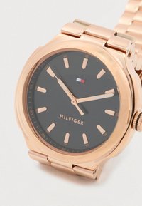 Rozā zelta rokas pulkstenis ar melnu ciparnīcu, zeltītām stundu atzīmēm un rādītājiem, kā arī Tommy Hilfiger logo uz ciparnīcas.