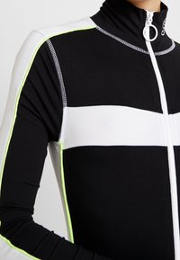 Svart och vit zip-up-jacka med hög krage, slim fit och reflekterande neongula detaljer längs sömmarna. Slät textur med stretch.