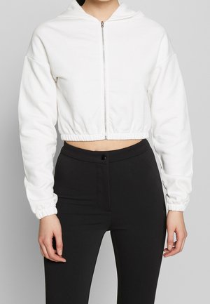 Mujer con sudadera blanca corta con cremallera y puños elásticos, y pantalones negros de talle alto, sobre un fondo liso.