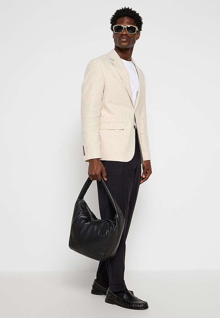 Beiger Leinenblazer, weißes T-Shirt, schwarze Hose und Loafers. Trägt eine große, weiche schwarze Schultertasche. Trägt übergroße Sonnenbrillen.