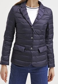 Kvinna i en marinblå kviltad kavaj med spetsiga lappar, knappstängning, fickor med klaff, över en vit t-shirt med rund halsringning och blå jeans.